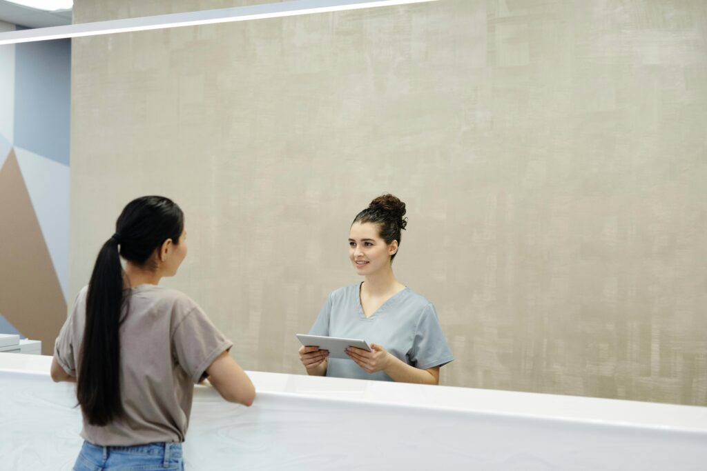 A smiling receptionist with a tablet assists a patient at a medical facility reception desk.